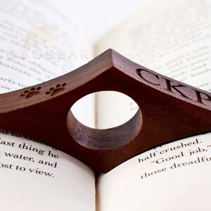 May include: A brown wooden book holder with a paw print design and the initials "CKP" engraved on the side. The holder is designed to hold a book open while reading.