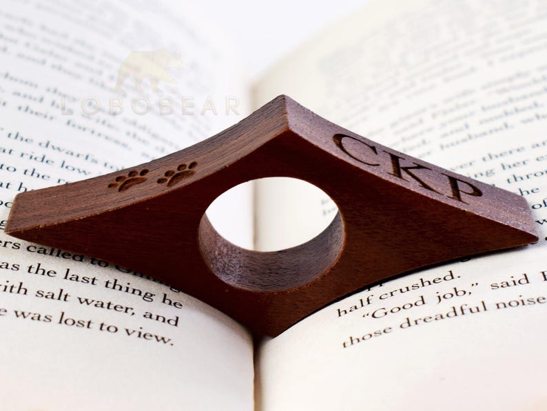 May include: A brown wooden book holder with a paw print design and the initials "CKP" engraved on the side. The holder is designed to hold a book open while reading.
