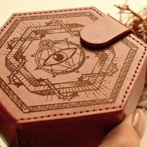 Puede incluir: Una caja hexagonal de cuero marrón rojizo con un diseño grabado decorativo que presenta un símbolo de ojo y patrones geométricos. La caja tiene un cierre de cuero a juego y está cosida con un hilo en contraste.