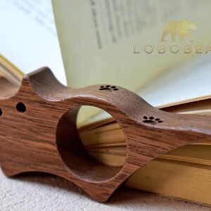 Puede incluir: Un sujetalibros de madera con un anillo para el dedo, con dise&ntilde;os de huellas de patas. El soporte de madera oscura est&aacute; colocado sobre las p&aacute;ginas de un libro abierto. La marca "LOBEBEAR" es visible.