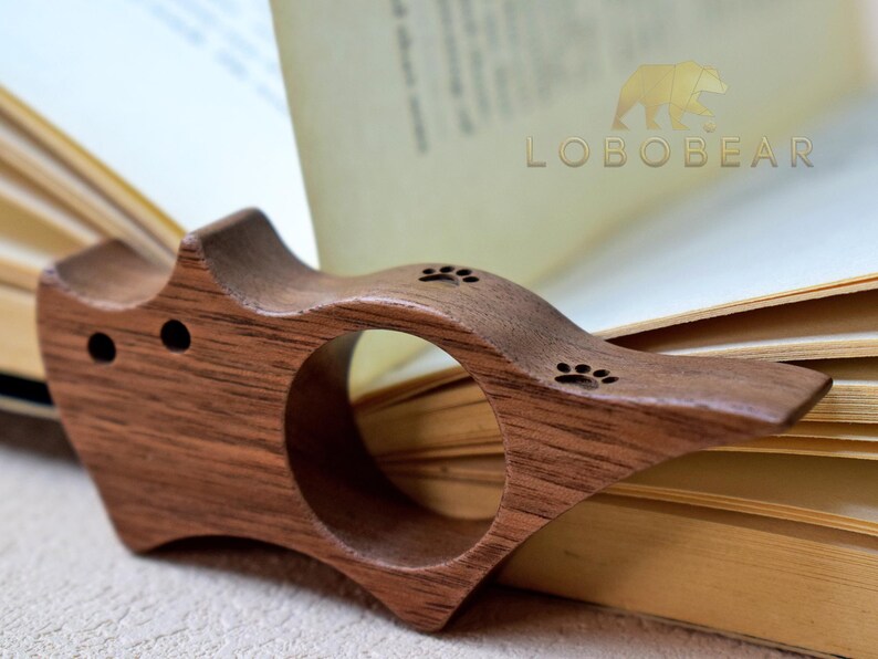 Puede incluir: Un sujetalibros de madera con un anillo para el dedo, con dise&ntilde;os de huellas de patas. El soporte de madera oscura est&aacute; colocado sobre las p&aacute;ginas de un libro abierto. La marca "LOBEBEAR" es visible.