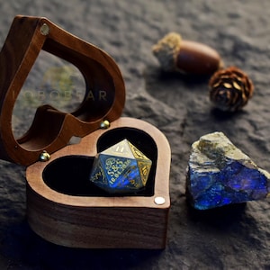 Peut inclure: Une boîte en bois en forme de cœur avec un couvercle transparent et une doublure noire contient un dé à multiples facettes gravé avec le texte S&S et 10.04.2026. Le dé présente des tons bleu et or irisés, accompagné d'une pierre brute et d'un gland.