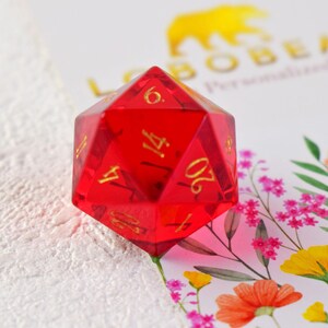 May include: A red, clear, 20-sided die with gold numbers. The die is sitting on a white surface with a floral pattern.