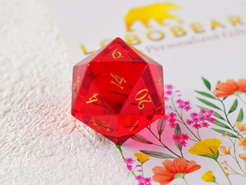 May include: A red, clear, 20-sided die with gold numbers. The die is sitting on a white surface with a floral pattern.