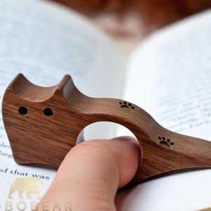 Puede incluir: Un marcap&aacute;ginas de madera con forma de gato con un anillo para el dedo. El marcap&aacute;ginas es de madera marr&oacute;n oscuro y presenta dos peque&ntilde;os ojos circulares y huellas de patas. El marcap&aacute;ginas est&aacute; colocado en un libro abierto.