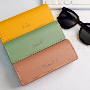 May include: Three personalised eyeglass cases in yellow, green, and peach. Each case has a name engraved on it: Lily, Gabrielle, and Hannah. Black sunglasses rest on the side.