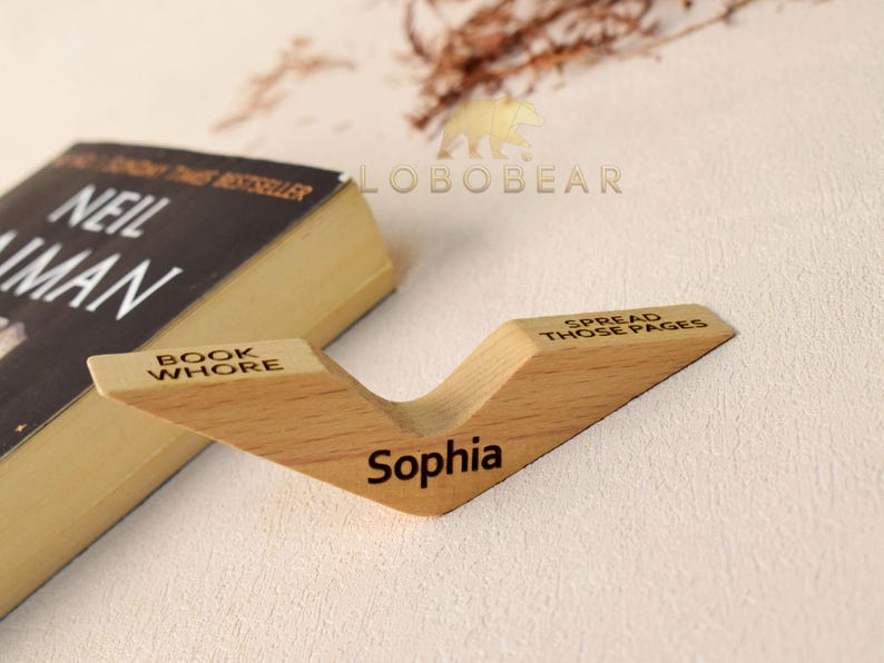 Peut inclure: Un support de livre en bois avec les mots "Book Whore", "Spread Those Pages" et "Sophia" grav&eacute;s dessus. Le support est brun clair et a une forme de V.