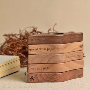 May include: A stack of five wooden thumb page holders with engraved text and symbols. The top piece reads BOOK WHORE, followed by pieces with the phrases spread those pages, just one more page, One more page, and a paw print and sun icon.