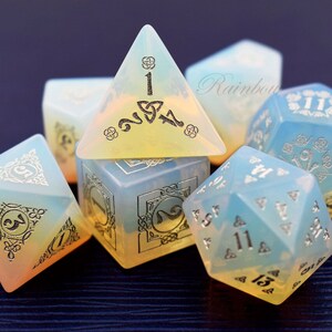 May include: A set of six iridescent dice with silver metallic numbers and intricate designs. The dice are made of a translucent, opalescent material that shifts color in the light. The dice are arranged in a cluster on a dark surface.