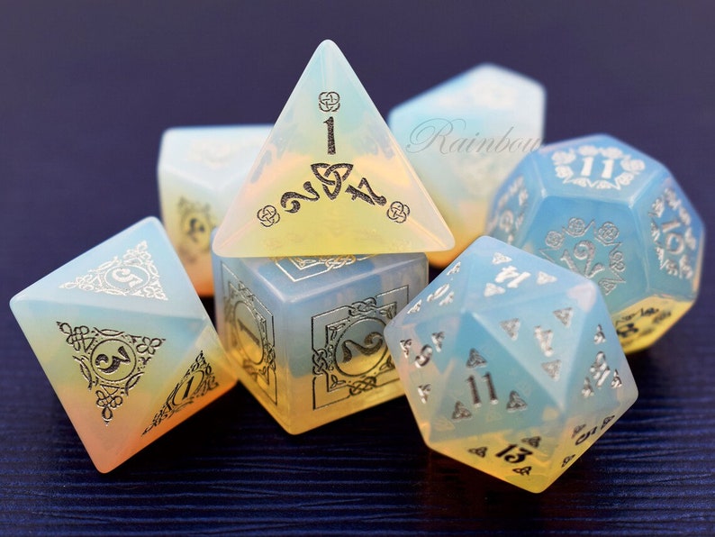 May include: A set of six iridescent dice with silver metallic numbers and intricate designs. The dice are made of a translucent, opalescent material that shifts color in the light. The dice are arranged in a cluster on a dark surface.