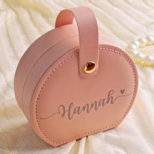 K&ouml;nnte beinhalten: Eine rosa, runde Schmuckschatulle mit Griff und dem Namen "Hannah" in Schreibschrift. Die Schatulle hat einen goldfarbenen Verschluss und ein gen&auml;htes Design. Ein Perlenarmband befindet sich im Hintergrund.