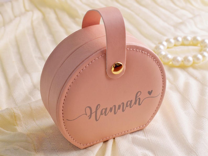 K&ouml;nnte beinhalten: Eine rosa, runde Schmuckschatulle mit Griff und dem Namen "Hannah" in Schreibschrift. Die Schatulle hat einen goldfarbenen Verschluss und ein gen&auml;htes Design. Ein Perlenarmband befindet sich im Hintergrund.