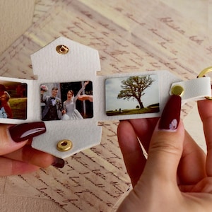 May include: A white leatherette photo keychain with three small photo inserts. The keychain has a gold-colored split ring and snap closure. The photos depict wedding scenes. The keychain is held open, displaying the photos.