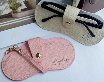 Puede incluir: Un estuche para gafas de color rosa claro texturizado con cierre de broche dorado, una correa de muñeca y el nombre Sophia impreso. Al lado hay un estuche color crema texturizado con gafas negras y las iniciales E|P impresas en el frente.