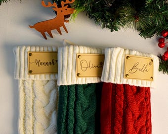 Op de afbeelding: Drie kabelgebreide kerstsokken in crème, groen en rood, elk met een gepersonaliseerd houten naamlabel. De namen "Hannah", "Olivia" en "Bella" worden weergegeven. Een houten rendier en feestelijke groenvoorziening maken de scène compleet.