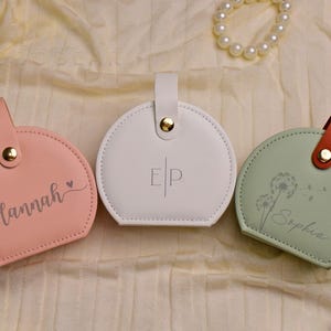 K&ouml;nnte beinhalten: Drei personalisierte runde Etuis in Rosa, Wei&szlig; und Gr&uuml;n. Das rosa Etui hat den Namen "Hannah" in Schreibschrift, das wei&szlig;e Etui hat die Initialen "EP" und das gr&uuml;ne Etui hat den Namen "Sophia" in Schreibschrift. Jedes Etui hat einen Griff.
