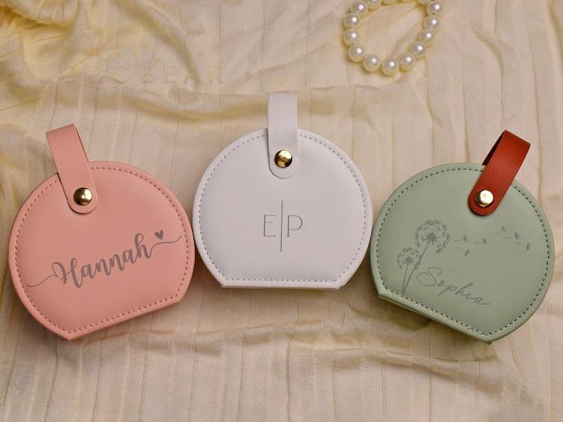 K&ouml;nnte beinhalten: Drei personalisierte runde Etuis in Rosa, Wei&szlig; und Gr&uuml;n. Das rosa Etui hat den Namen "Hannah" in Schreibschrift, das wei&szlig;e Etui hat die Initialen "EP" und das gr&uuml;ne Etui hat den Namen "Sophia" in Schreibschrift. Jedes Etui hat einen Griff.
