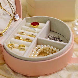 K&ouml;nnte beinhalten: Eine rosa, runde Schmuckschatulle mit mehreren F&auml;chern, gef&uuml;llt mit Halsketten, Ringen und Armb&auml;ndern. Der Schmuck umfasst goldene, silberne und Perlen-Akzente. Eine wei&szlig;e Schachtel befindet sich im Hintergrund, und eine Perlenkette ist rechts.