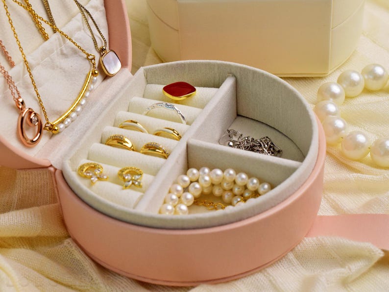 K&ouml;nnte beinhalten: Eine rosa, runde Schmuckschatulle mit mehreren F&auml;chern, gef&uuml;llt mit Halsketten, Ringen und Armb&auml;ndern. Der Schmuck umfasst goldene, silberne und Perlen-Akzente. Eine wei&szlig;e Schachtel befindet sich im Hintergrund, und eine Perlenkette ist rechts.
