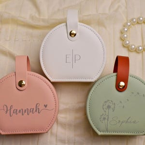 K&ouml;nnte beinhalten: Drei runde, personalisierte Lederbeutel in Pastellfarben: Rosa, Wei&szlig; und Gr&uuml;n. Jeder Beutel hat einen Griff, einen goldfarbenen Druckknopfverschluss und einen Namen oder Initialen auf der Vorderseite. Ein Perlenarmband ist ebenfalls sichtbar.