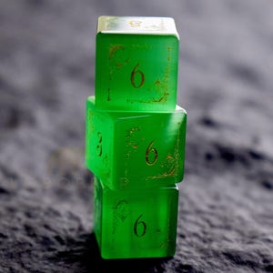 Peut inclure: Trois d&eacute;s verts translucides empil&eacute;s, chacun affichant le chiffre six. Les d&eacute;s pr&eacute;sentent des motifs d&eacute;coratifs dor&eacute;s autour des bords des chiffres. Les d&eacute;s sont en forme de cube et semblent &ecirc;tre faits d'un mat&eacute;riau solide.