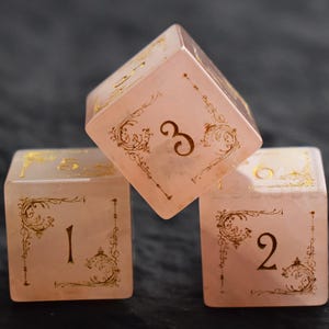 Op de afbeelding: Drie roze dobbelstenen met goudkleurige cijfers en decoratieve krullen. De dobbelstenen zijn in een piramidevorm gerangschikt, met de cijfers 1, 2 en 3 zichtbaar. De dobbelstenen lijken van een doorschijnend materiaal te zijn.