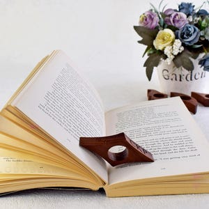 May include: A wooden book holder in the shape of a bookmark, with a dark brown finish, is holding open a book with white pages. The book holder is resting on a white surface. The book has the text "The Bachelor Games" printed on the bottom page.