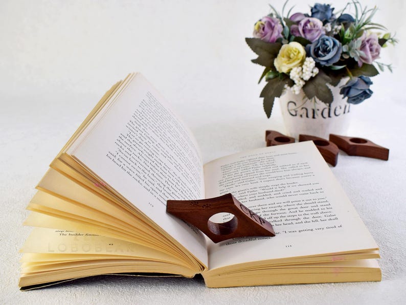 May include: A wooden book holder in the shape of a bookmark, with a dark brown finish, is holding open a book with white pages. The book holder is resting on a white surface. The book has the text "The Bachelor Games" printed on the bottom page.