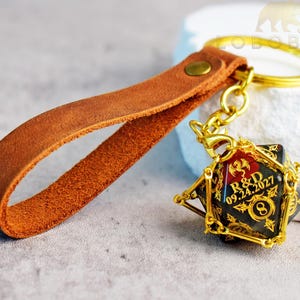 Puede incluir: Una correa de llavero de cuero marrón unida a un anillo de metal dorado y un dado negro de múltiples caras encerrado en una jaula de metal dorado. El dado presenta imágenes de dragones dorados y el texto R&D 09.24.2027 con un símbolo de infinito.
