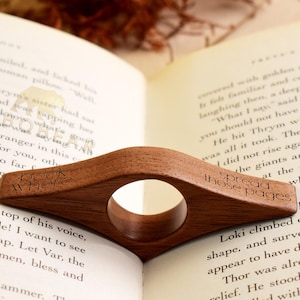 May include: A wooden book page holder with a circular opening, resting on an open book. The holder is engraved with the words "book where" and "spread those pages". The wood has a warm, brown tone.