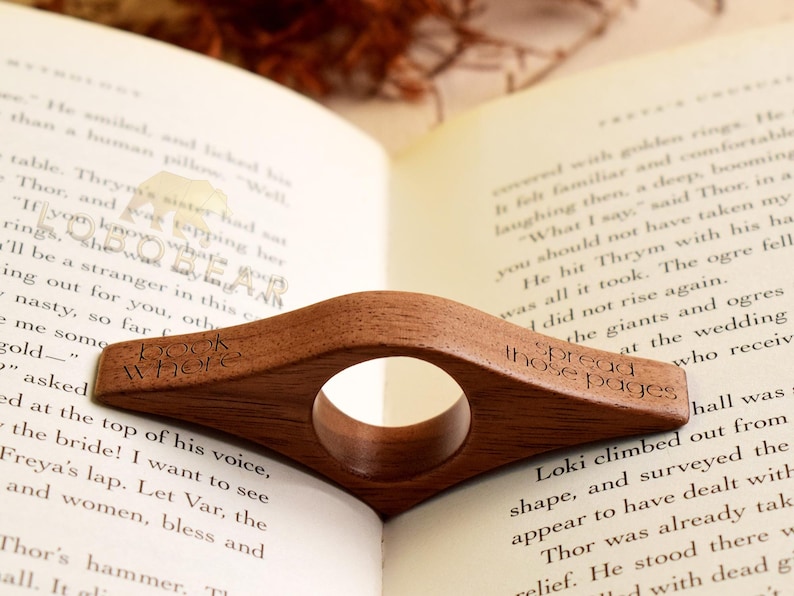 May include: A wooden book page holder with a circular opening, resting on an open book. The holder is engraved with the words "book where" and "spread those pages". The wood has a warm, brown tone.