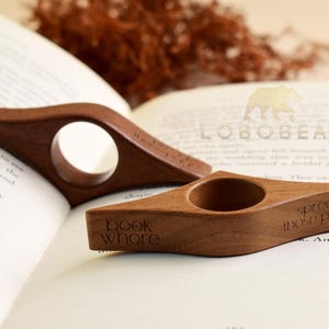 May include: Two wooden book holders with a hole for the thumb, resting on an open book. One has the text "book whore" and the other has "spread those pages". The holders are a warm brown color.