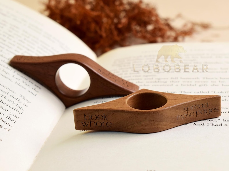 May include: Two wooden book holders with a hole for the thumb, resting on an open book. One has the text "book whore" and the other has "spread those pages". The holders are a warm brown color.
