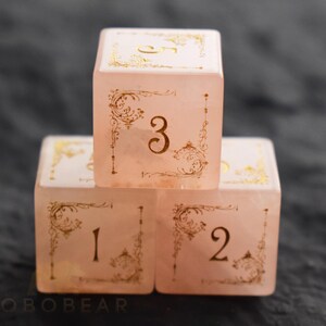 Peut inclure: Trois d&eacute;s roses avec des filigranes dor&eacute;s et les chiffres 1, 2, 3 et 5. Les d&eacute;s sont empil&eacute;s, le chiffre 3 face au spectateur. Les d&eacute;s ont un aspect translucide et sont pos&eacute;s sur un fond sombre.