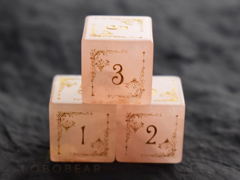 Peut inclure: Trois d&eacute;s roses avec des filigranes dor&eacute;s et les chiffres 1, 2, 3 et 5. Les d&eacute;s sont empil&eacute;s, le chiffre 3 face au spectateur. Les d&eacute;s ont un aspect translucide et sont pos&eacute;s sur un fond sombre.
