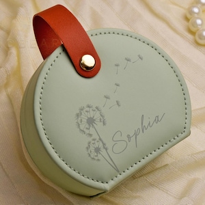 K&ouml;nnte beinhalten: Eine hellgr&uuml;ne, herzf&ouml;rmige Schmuckschatulle mit einer gen&auml;hten Bord&uuml;re und einem orangefarbenen Griff. Die Schatulle zeigt ein Pusteblumen-Design und den Namen "Sophia" in Schreibschrift. Ein goldener Knopf sichert den Griff.