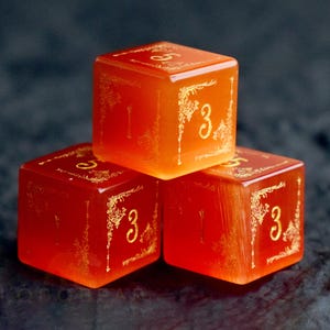 Peut inclure: Trois d&eacute;s orange avec des filigranes dor&eacute;s et le chiffre 3 sur plusieurs faces. Les d&eacute;s sont empil&eacute;s, un d&eacute; sur les deux autres. Les d&eacute;s sont translucides et brillent l&eacute;g&egrave;rement.