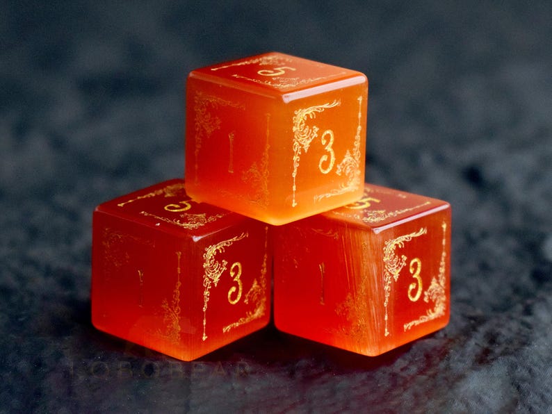Peut inclure: Trois d&eacute;s orange avec des filigranes dor&eacute;s et le chiffre 3 sur plusieurs faces. Les d&eacute;s sont empil&eacute;s, un d&eacute; sur les deux autres. Les d&eacute;s sont translucides et brillent l&eacute;g&egrave;rement.