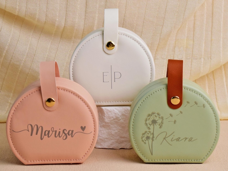 K&ouml;nnte beinhalten: Drei runde, personalisierte Kosmetiktaschen in Pastellfarben. Die Taschen haben Griffe und goldfarbene Hardware. Eine ist pfirsichfarben mit "Marisa" und einem Herz, eine andere ist wei&szlig; mit "EP", und die dritte ist gr&uuml;n mit "Kiara" und einem L&ouml;wenzahnmotiv.
