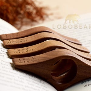 May include: A stack of wooden book holders, each engraved with "book whore", rests on an open book. The holders are a warm brown color and have a curved design with a finger hole. The background includes a logo and dried foliage.