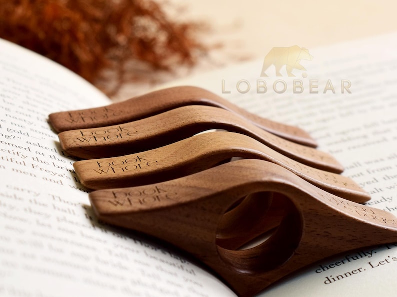 May include: A stack of wooden book holders, each engraved with "book whore", rests on an open book. The holders are a warm brown color and have a curved design with a finger hole. The background includes a logo and dried foliage.