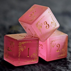 Peut inclure: Trois d&eacute;s roses translucides avec des chiffres dor&eacute;s et des motifs d&eacute;coratifs. Les d&eacute;s sont empil&eacute;s, avec les chiffres 1, 2, 3, 4 et 5 visibles. Les d&eacute;s sont pour les jeux de soci&eacute;t&eacute;.
