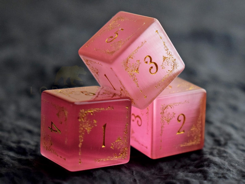 Peut inclure: Trois d&eacute;s roses translucides avec des chiffres dor&eacute;s et des motifs d&eacute;coratifs. Les d&eacute;s sont empil&eacute;s, avec les chiffres 1, 2, 3, 4 et 5 visibles. Les d&eacute;s sont pour les jeux de soci&eacute;t&eacute;.