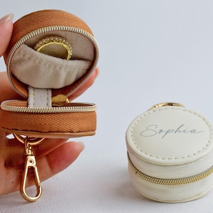 May include: A round, tan zippered jewelry case with a gold-tone metal clasp and a beige interior holding a ring. Beside it sits a closed, round, white zippered jewelry case with the name Sophia printed in a cursive font on the top surface.