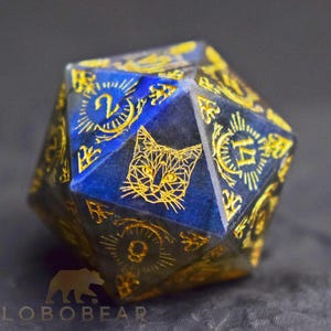 Peut inclure: Un dé à vingt faces en pierre bleu foncé avec des chiffres gravés de couleur dorée et une illustration géométrique de tête de chat. Le dé présente des motifs de rayons de soleil autour des chiffres et le texte LOBOBEAR sur le bord inférieur.