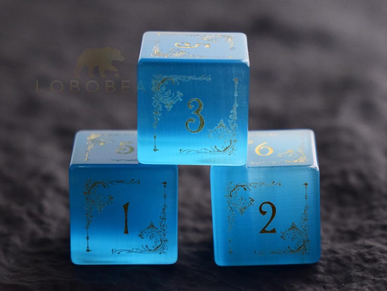 Peut inclure: Trois d&eacute;s bleus translucides avec des chiffres dor&eacute;s et des ornements d&eacute;coratifs. Les d&eacute;s sont num&eacute;rot&eacute;s 1, 2, 3, 5 et 6. Les d&eacute;s sont empil&eacute;s les uns sur les autres.