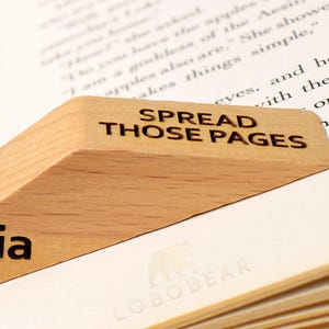 Peut inclure: Un serre-livres en bois avec le texte "SPREAD THOSE PAGES" et le nom "Sophia" grav&eacute; dessus. Le serre-livres est pos&eacute; sur un livre avec le texte "LOBOBEAR" imprim&eacute; sur la page.