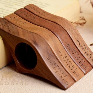May include: Four wooden book holders in varying shades of brown, each with a curved design and a finger hole. The text "BOOK WHOLE" and "spread those pages" is engraved on the holders. The items are resting on a book.