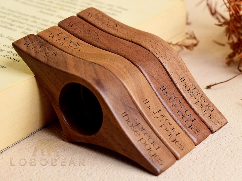 May include: Four wooden book holders in varying shades of brown, each with a curved design and a finger hole. The text "BOOK WHOLE" and "spread those pages" is engraved on the holders. The items are resting on a book.