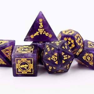 May include: Set of six purple and gold dice with Celtic knot designs. The dice are made of amethyst and have gold numbers and symbols.